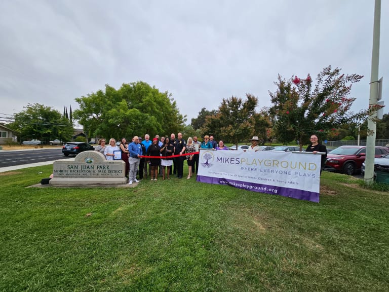 Group of people holding a Mikes Playground sign and a red ribbon